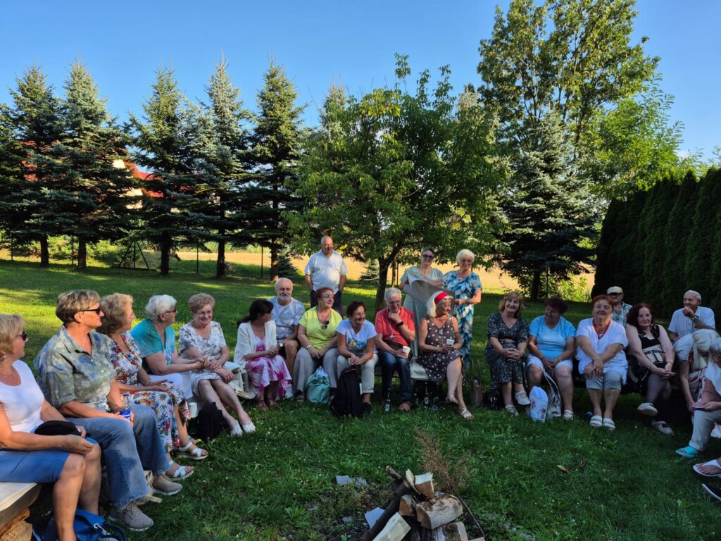 Grupa osób siedzi w kręgu na trawie podczas plenerowego spotkania w ogrodzie. Uczestnicy rozmawiają w otoczeniu drzew, a pośrodku przygotowane jest niewielkie ognisko.