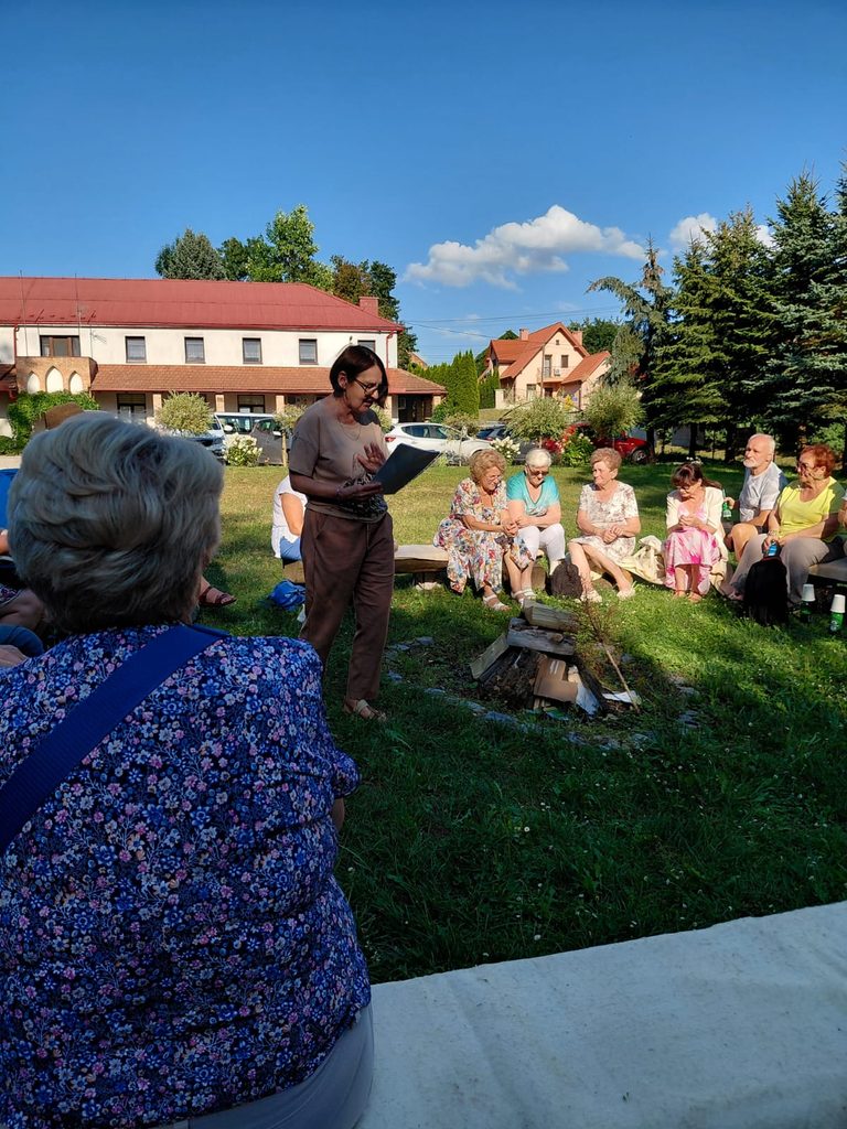 Uczestnicy Kolpingowskiego Spotkania Wolontariuszy siedzą w kręgu na trawie wokół przygotowanego ogniska, a prowadząca stojąca pośrodku czyta materiały związane ze spotkaniem.
