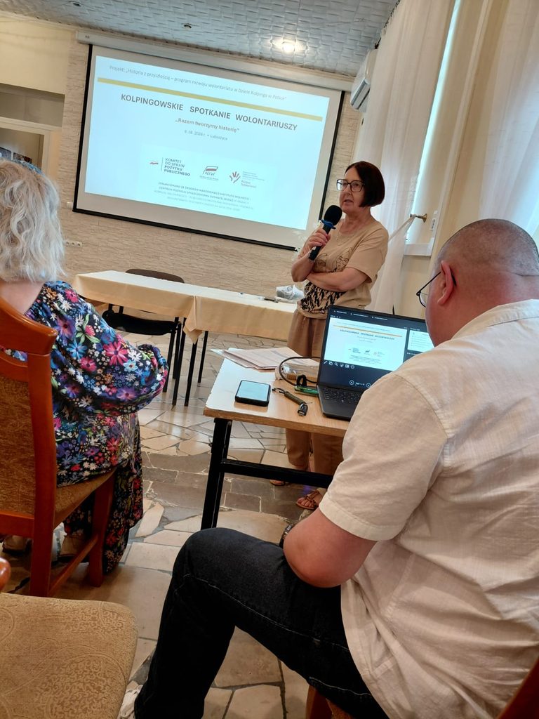 Prowadząca z mikrofonem przemawia do uczestników Kolpingowskiego Spotkania Wolontariuszy. W tle na ekranie wyświetlona jest prezentacja dotycząca wydarzenia i projektu „Historia z przyszłością”.