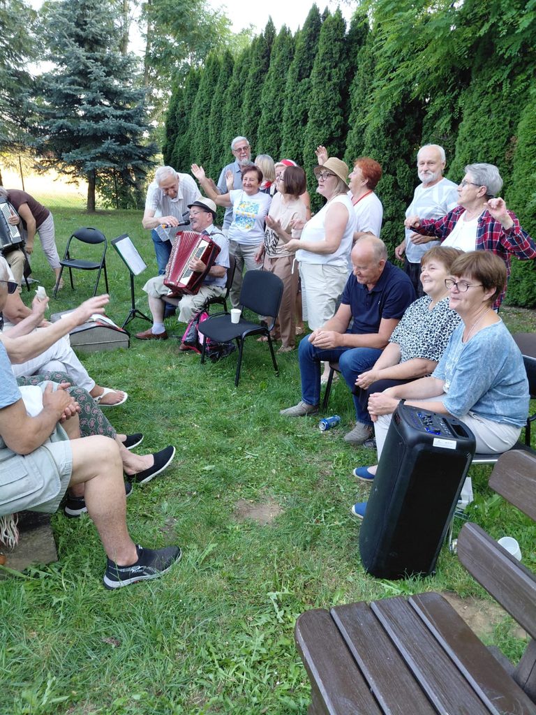 Uczestnicy spotkania wspólnie śpiewają w ogrodzie przy akompaniamencie akordeonu. Grupa siedzi i stoi w kręgu, tworząc radosną, integracyjną atmosferę.