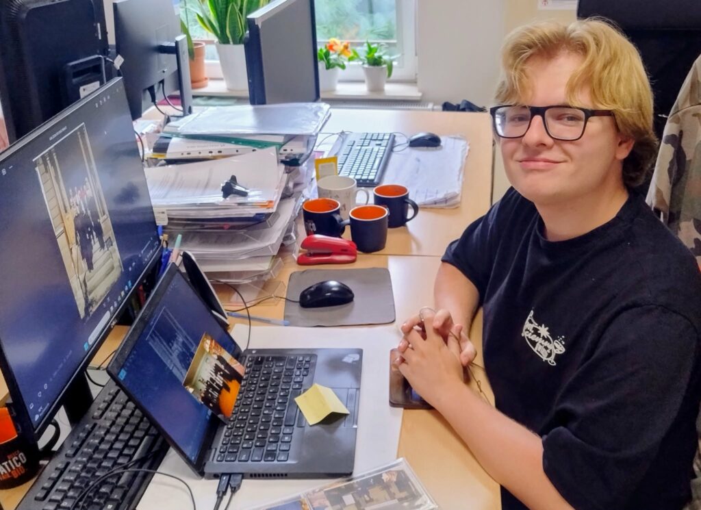 Młody mężczyzna w okularach siedzi przy biurku w biurze, obok laptopa i dwóch monitorów. Na biurku znajdują się dokumenty, kubki i akcesoria biurowe, a w tle widać okno z roślinami.