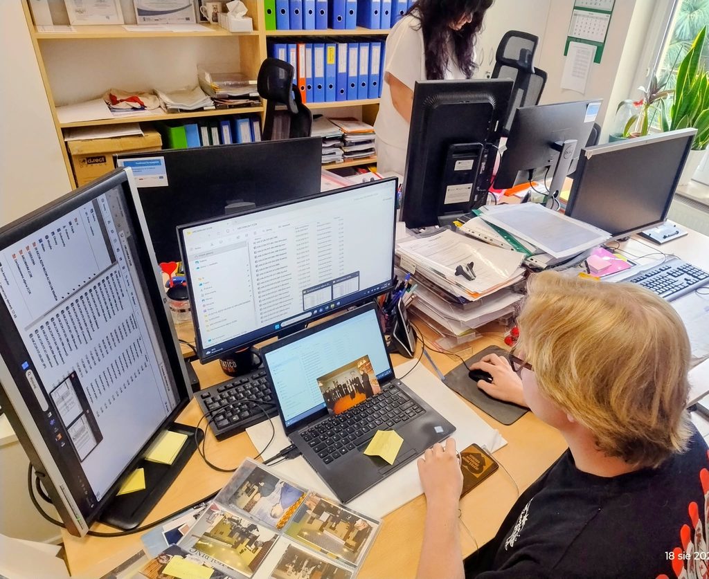 Osoba siedząca przy biurku przegląda i digitalizuje archiwalne fotografie. Zdjęcia są wyświetlane na laptopie i monitorze, a obok znajdują się albumy ze zdjęciami oraz stosy dokumentów.