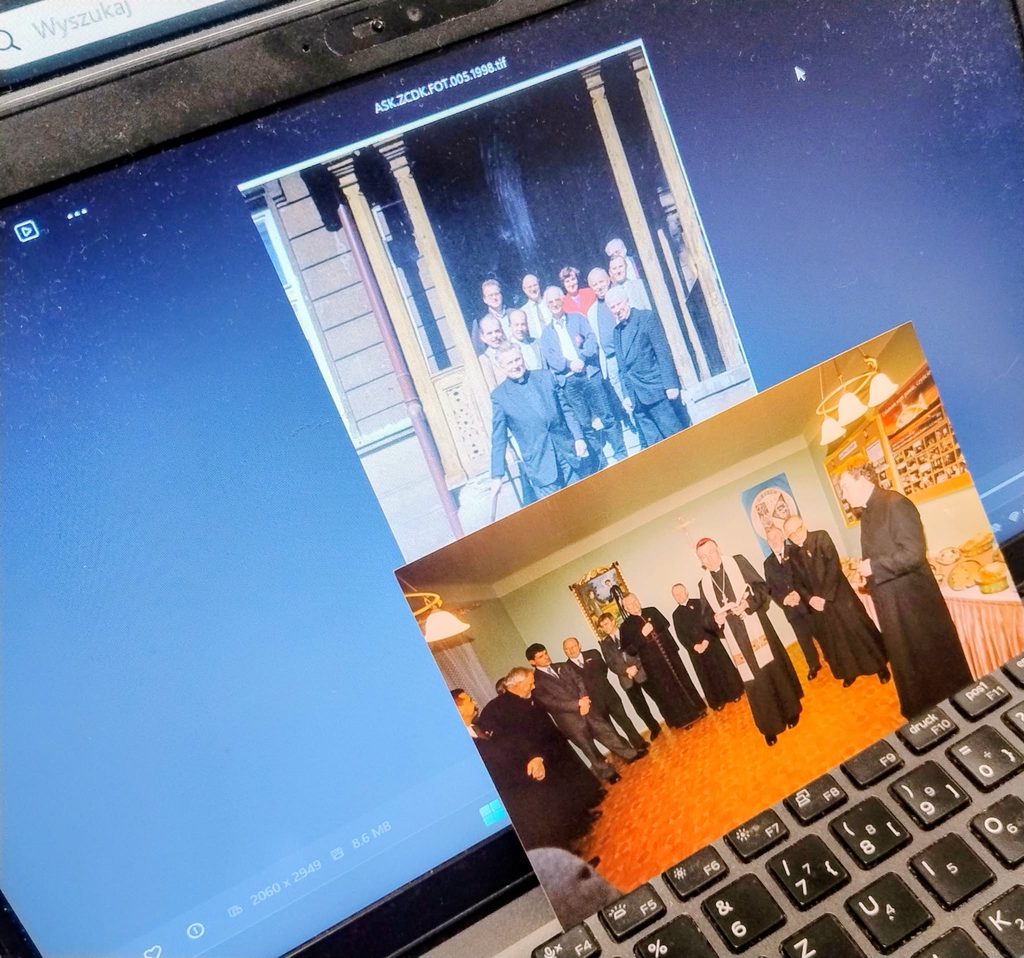 Zbliżenie na laptop, na którego ekranie wyświetlane jest archiwalne zdjęcie grupowe; na klawiaturze leży odbitka starej fotografii przedstawiającej grupę osób podczas uroczystości.