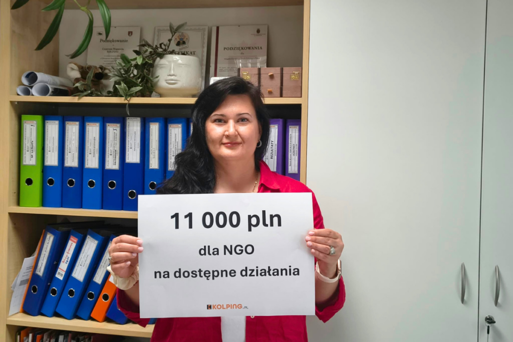Kobieta trzyma kartkę z napisem „11 000 PLN dla NGO na dostępne działania” i logo KOLPING.PL. W tle regał z dokumentami i segregatorami.
